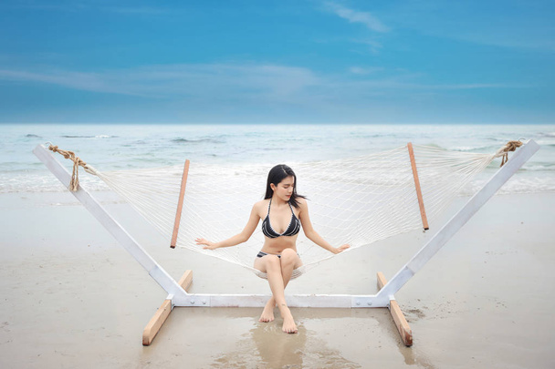 Sexy asijská žena v černých a bílých bikinách, které lžou nebo sedí na houpací síti na písečné pláži s krásnou tváří a modrou oblohou na dovolené v letním čase - Fotografie, Obrázek