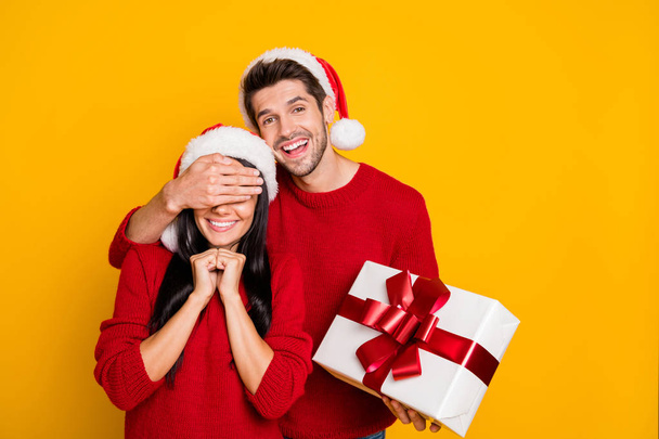 Portrait of charming guy in xmas cap hide his lovely sweethearts eyes give her package wear red sweater isolated over yellow background - Foto, Bild