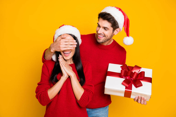 Portrait of handsome man with brown hair hide his excited wife eyes give her package wear red pullover isolated over yellow background - Fotó, kép