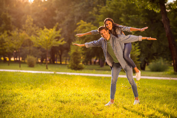 Full body photo of charming married people with brunet hair raising hands piggyback wearing denim jeans jacket blazers outside in park - Photo, image