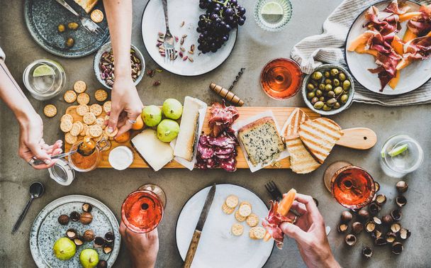 Mid-summer picnic with wine and snacks. Flat-lay of charcuterie and cheese board, rose wine, nuts, olives and peoples hands with snacks over concrete table background, top view - Foto, immagini