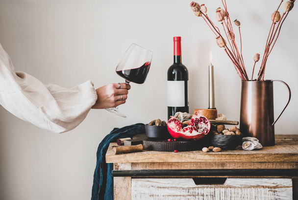 Red wine, board with cheese, fruit, almonds and olives, candles, decorative flowers on kitchen counter and womans hand holding glass of wine, white wall at background. Wine tasting concept - Photo, image