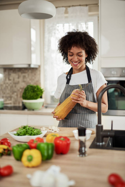 Beautiful mixed race housewife in apron standing in kitchen and holding zucchini On kitchen counter are vegetables. Meal preparation. - Фото, изображение