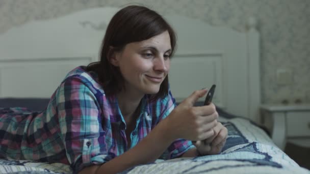 Attractive young female lying on the bed and surfing online via smartphone and laptop  - Imágenes, Vídeo