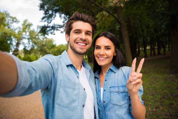 Close up photo of charming couple having brunette hair making photos v-signs wearing denim jeans shirt outside in park - Foto, Imagem