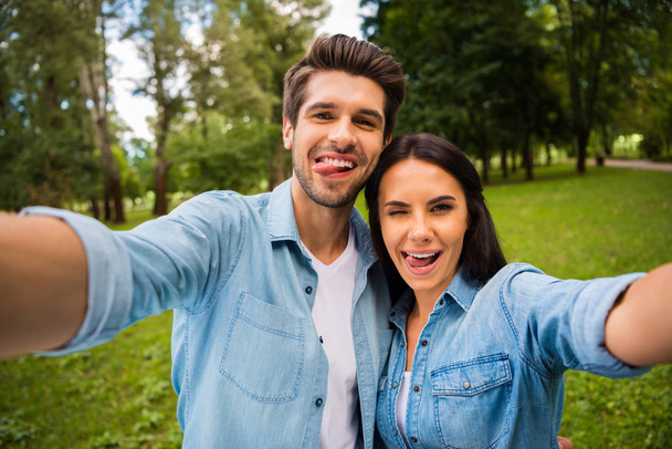 Close up photo of childish spouses with brunet hair making photo showing their tongues winking blinking wearing denim jeans shirt outdoors - Foto, imagen