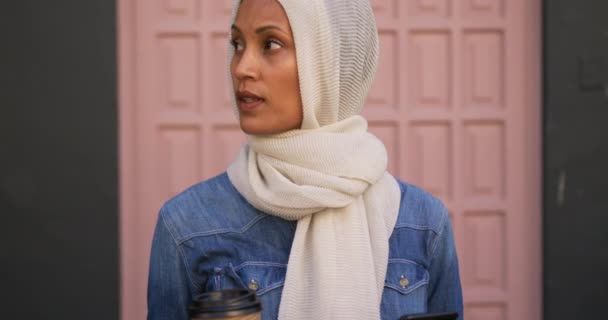 Front view of a young mixed race woman wearing a hijab standing in a city street, using a smartphone holding a coffee cup and smiling to camera - 映像、動画
