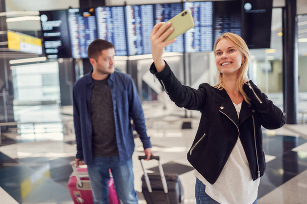 Image of man carrying luggage and woman taking selfie walking in waiting room at airport - Foto, imagen