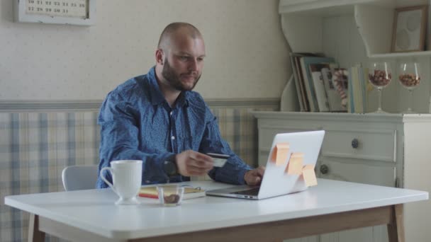 Young male freelance making purchase via credit card and working at laptop sitting at the table with tea and snacks  - Filmagem, Vídeo