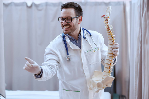 Joven perspectiva sonriente médico caucásico en uniforme blanco y con estetoscopio alrededor del cuello sosteniendo la columna vertebral y apuntando con el modelo de dedo mientras está de pie en el hospital
. - Foto, imagen