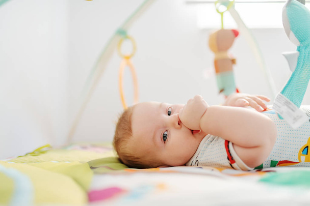 Widok z boku piękny sześciomiesięczny chłopiec w kolorowe body patrząc w aparat i ssać kciuk leżąc w łóżku otoczony zabawkami. - Zdjęcie, obraz
