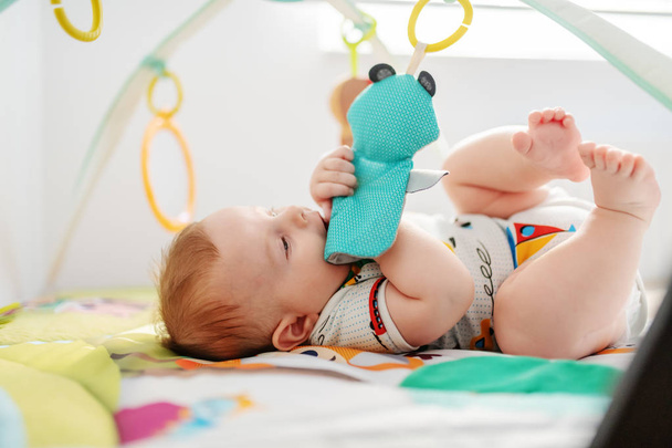 Sevimli altı aylık çocuk yatakta uzanıyor, ısırıyor ve beşik oyuncaklarıyla oynuyor. Sağlıklı yetiştirme kavramı. - Fotoğraf, Görsel