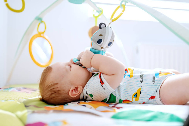 Adorable six months old boy lying in bed biting and playing with crib toys. Healthy upbringing concept. - Photo, Image