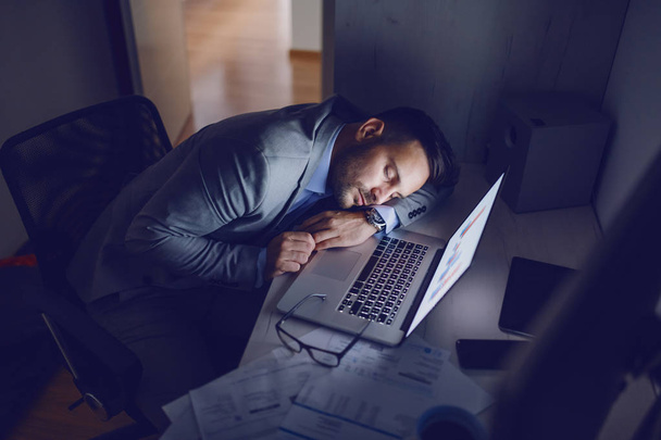 Overwerkte toegewijde hardwerkende werknemer in grijs pak die 's nachts op kantoor slaapt. Op het bureau zijn rapporten, brillen en laptop. - Foto, afbeelding
