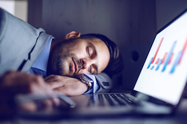 Overwerkte toegewijde hardwerkende werknemer in grijs pak die 's nachts op kantoor slaapt. Op het bureau zijn rapporten en laptop. - Foto, afbeelding