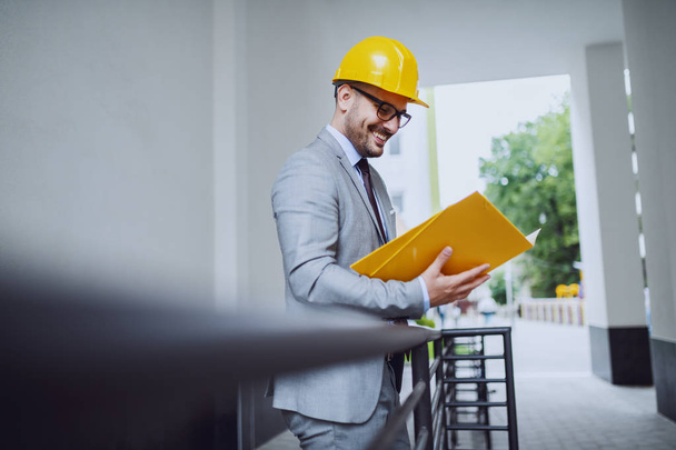 Vista lateral de um arquiteto caucasiano sorridente focado em terno e com capacete e óculos olhando para documentos na pasta ao ar livre
. - Foto, Imagem