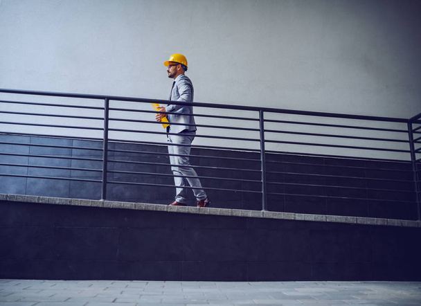 A side view of a businessman in a grey suit and safety gear on his head walking upstairs along the rail - Photo, Image