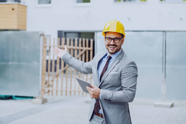 Guapo caucásico feliz arquitecto en traje gris y con casco amarillo en la cabeza que sostiene la tableta y muestra el sitio de construcción
. - Foto, Imagen