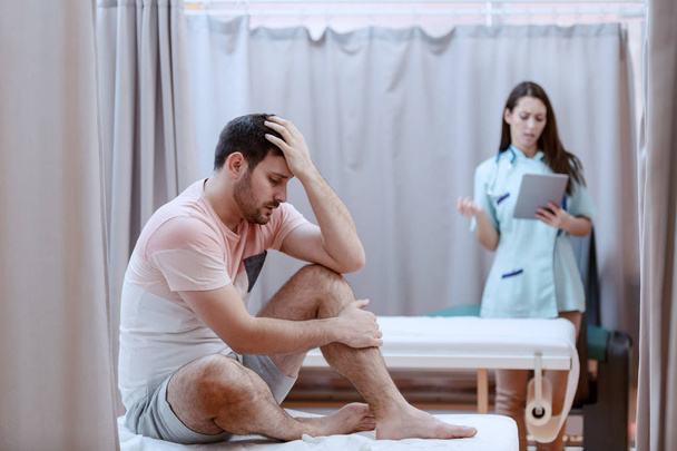 junge kaukasische männliche Patientin, die auf dem Krankenhausbett sitzt und Schmerzen fühlt. im Hintergrund Krankenschwester mit Tablette. - Foto, Bild