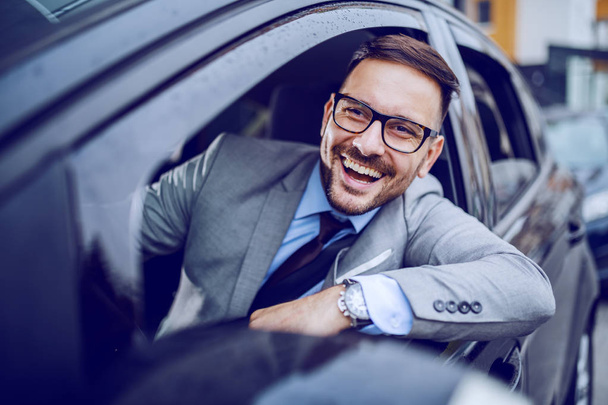 Χαμογελώντας επιχειρηματίας κοιτάζοντας κάτω παράθυρο, ενώ η οδήγηση ακριβό αυτοκίνητό του. Σχέδιο επαγγελματικού ταξιδιού. - Φωτογραφία, εικόνα