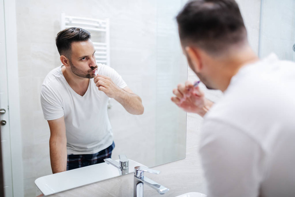 Handsome caucasian man standing in bathroom in pajamas and washing his teeth in the morning and watching himself in the mirror. - Фото, зображення