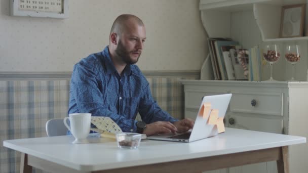 Young male freelance working at laptop and smartphone sitting at the table with tea and snacks  - Záběry, video