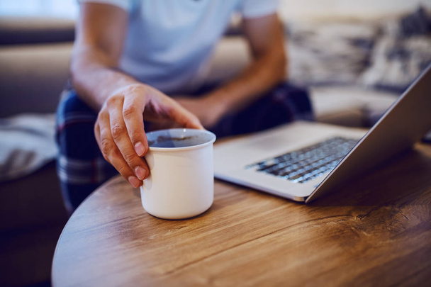 Close up of man taking mug with morning coffee from desk. Man is dressed in pajamas, sitting on sofa in living room and using laptop. Weekend activities. Selective focus on hand. - Фото, зображення