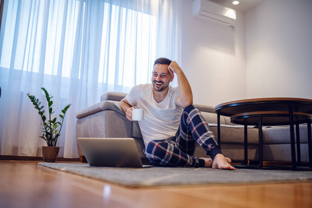 Charismatic smiling caucasian man in pajamas sitting on the floor in living room, leaning on sofa, holding mug with coffee and looking at laptop. Weekend activities concept. - 写真・画像