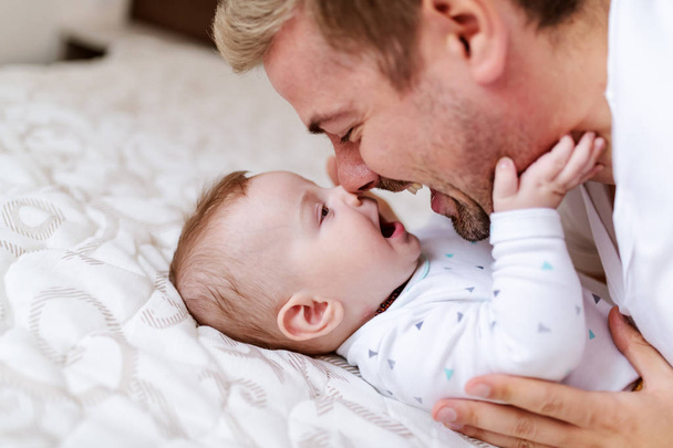 Sluiten van schattige zes maanden oude baby jongen liggend op bed en spelen met zijn vader. Papa praat met zoon en knuffelt hem.. - Foto, afbeelding