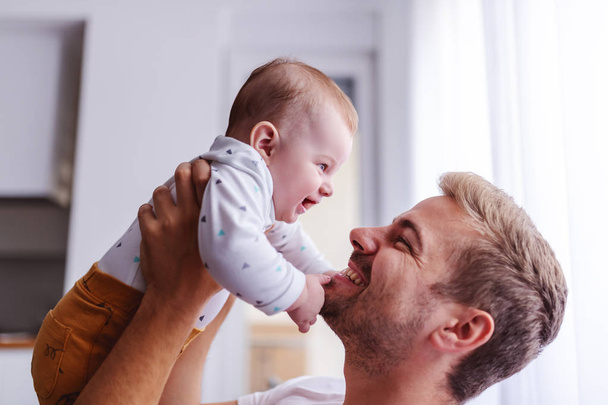 Happy smiling handsome young caucasian dad lifting his loving 6 months old son while standing in living room next to window. Baby is touching dad's face and laughing. Unconditional love concept. - Photo, image