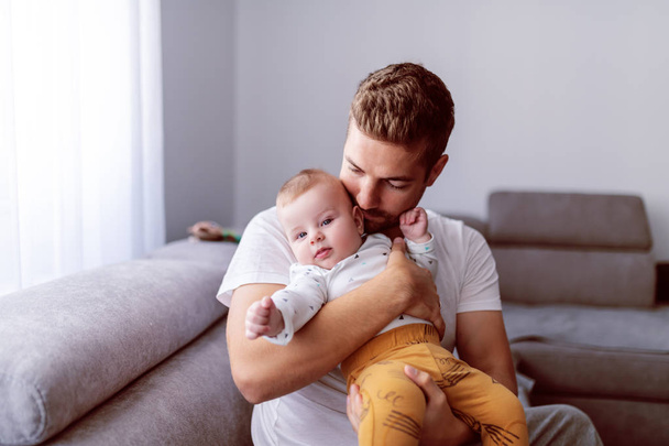 Yakışıklı, beyaz, genç bir baba, sevgi dolu 6 aylık oğlunu oturma odasında kanepede otururken tutuyor. Bebek kameraya bakıyor. Aile zamanı kavramı. - Fotoğraf, Görsel