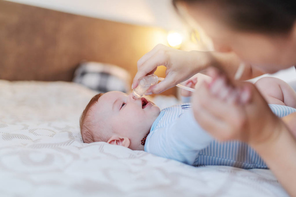 Zbliżenie na opiekuńczą białą matkę używającą pompki do nosa do czyszczenia nosa dziecka. Dziecko leżące na łóżku ubrane w body suit. - Zdjęcie, obraz