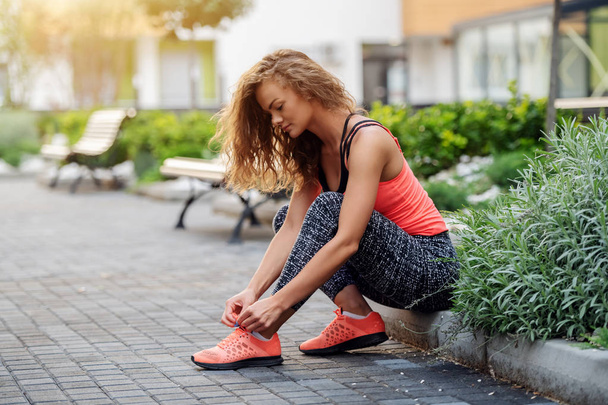 Charming fit caucasian woman dressed in sports wear sitting on roadside and tying shoelace. Park exterior. - Fotoğraf, Görsel
