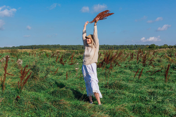 Happy beautiful blond woman walking in a green field with a bouquete of dry brown plants - Fotoğraf, Görsel