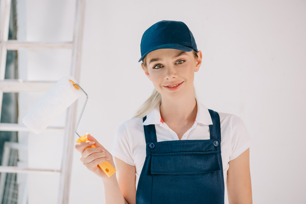 beautiful young woman in uniform holding paint roller and smiling at camera - 写真・画像