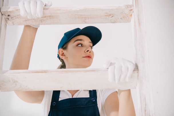 pretty young painter in uniform climbing on ladder and looking away - Photo, Image