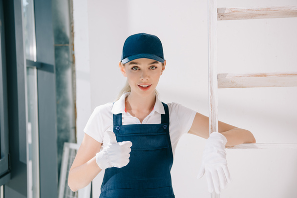 pretty young painter in uniform showing thumb up while looking at camera - Foto, Imagem