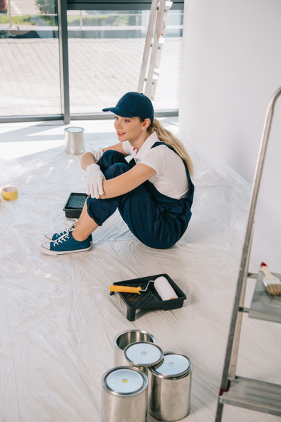 beautiful young painter in overalls and cap sitting on floor near paint roller in roller tray and cans with paint - Fotoğraf, Görsel