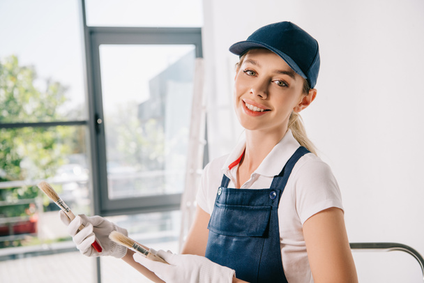 attractive young painter in uniform holding paintbrushes and smiling at camera - Foto, imagen