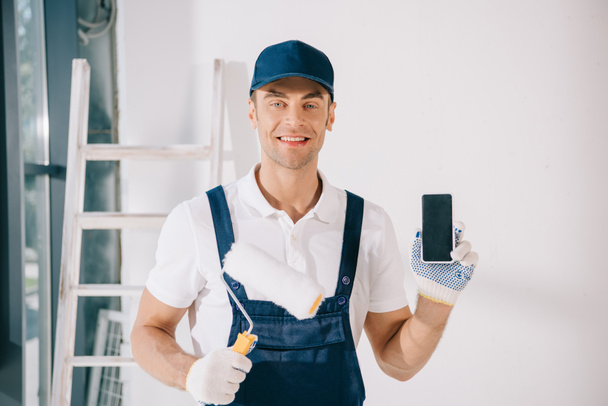 joven pintor guapo en uniforme sosteniendo rodillo de pintura y mostrando teléfono inteligente con pantalla en blanco
 - Foto, imagen