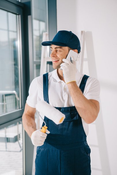 handsome young painter in uniform looking away while holding paint roller and talking on smartphone - Photo, Image