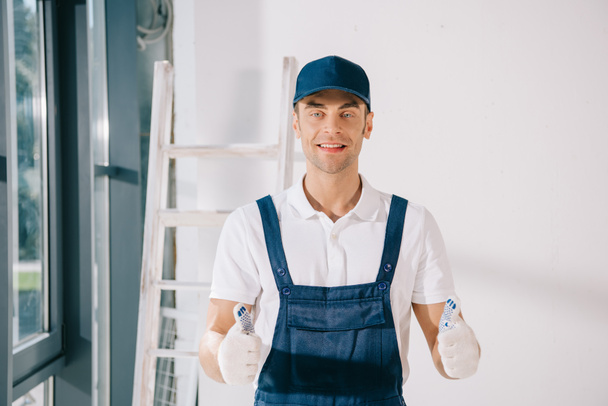 handsome painter in uniform showing thumbs up and smiling at camera - Photo, Image