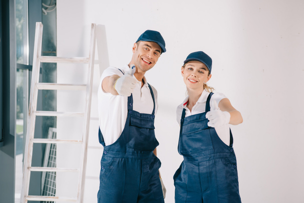 young painters in overalls showing thumbs up while smiling at camera - Photo, Image