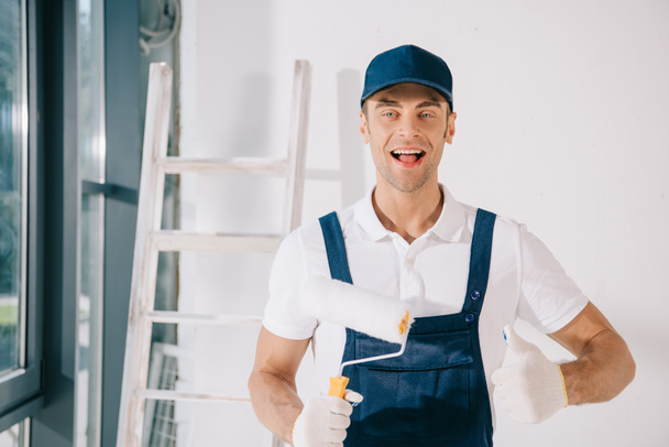 handsome young painter in uniform holding paint roller, showing thumb up and smiling at camera - Foto, Imagen