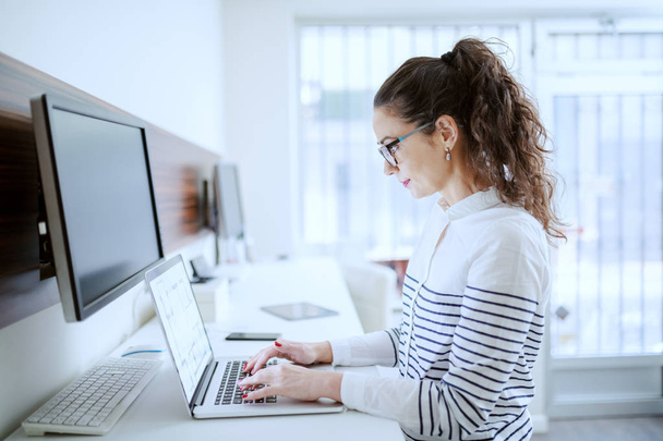 Side view of hard working Caucasian businesswoman with brown curly hair and eyeglasses typing on laptop. Office interior. - Fotó, kép