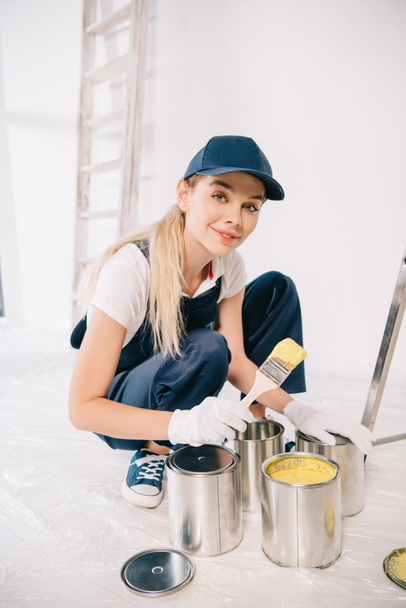 beautiful young painter in overalls and cap holding paintbrush near can with yellow paint - Photo, Image