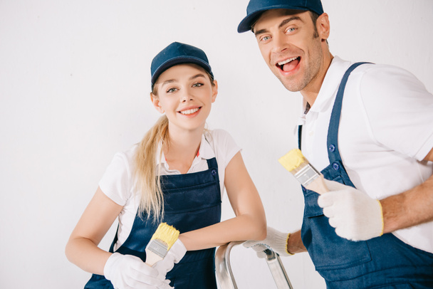 two cheerful painters in uniform holding paintbrushes and smiling at camera - Photo, Image