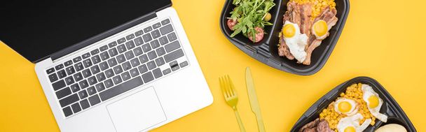 panoramisch shot van eco-pakketten met groenten, vlees, gebakken eieren en salade, laptop, plastic vork en mes geïsoleerd op geel     - Foto, afbeelding