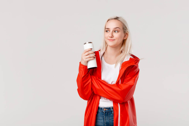 Pretty blond woman in a red raincoat holding white thermal mug with hot beverage isolated over white background. - Foto, immagini