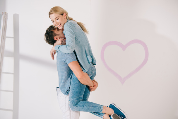 happy young man holding girlfriend on hands near pink heart drawn on white wall - Foto, afbeelding